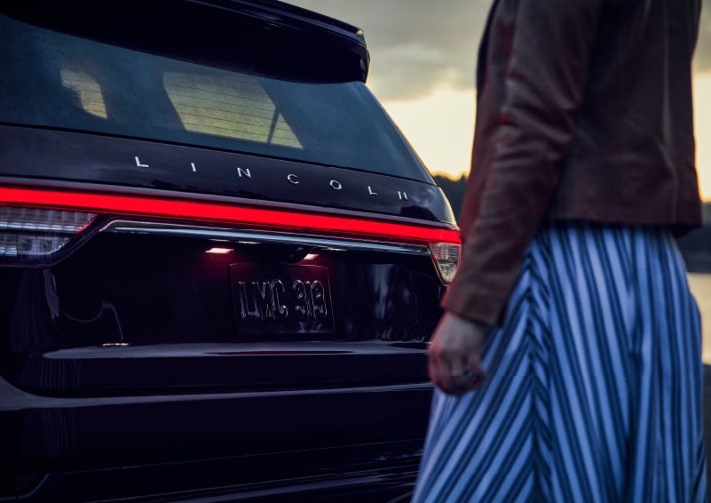 A person is shown near the rear of a 2024 Lincoln Aviator® SUV as the Lincoln Embrace illuminates the rear lights | Lincoln Demo 4 in Derwood MD