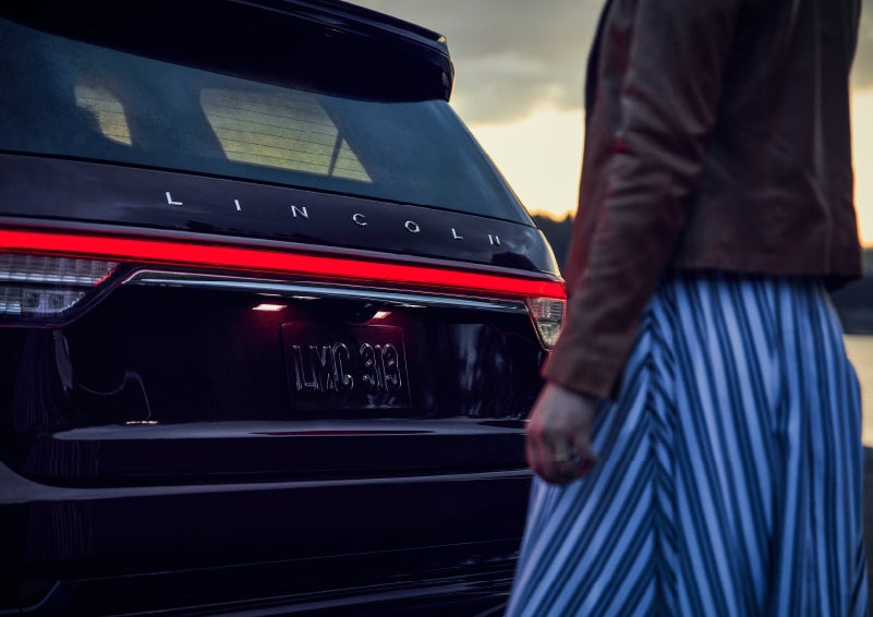A person is shown near the rear of a 2024 Lincoln Aviator® SUV as the Lincoln Embrace illuminates the rear lights | Lincoln Demo 4 in Wooster OH
