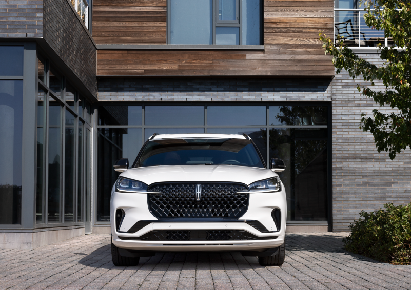 A white 2025 Lincoln Aviator® SUV is shown parked outside a modern building with gray brick and wood accents. | Lincoln Demo 4 in Derwood MD