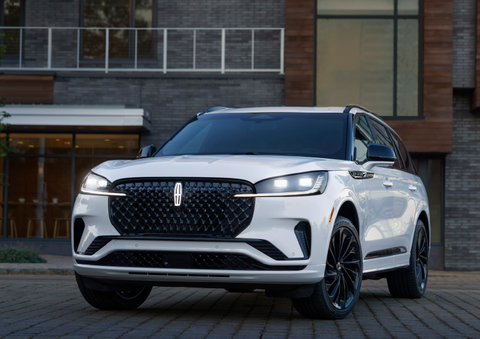 A white 2025 Lincoln Aviator® SUV is shown parked outside a modern building with gray brick and wood accents. | Lincoln Demo 4 in Derwood MD